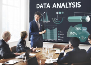 Business professional presenting data analytics dashboard to executive team in modern conference room, with large screen displaying charts, graphs, and performance metrics including percentages and trend analysis