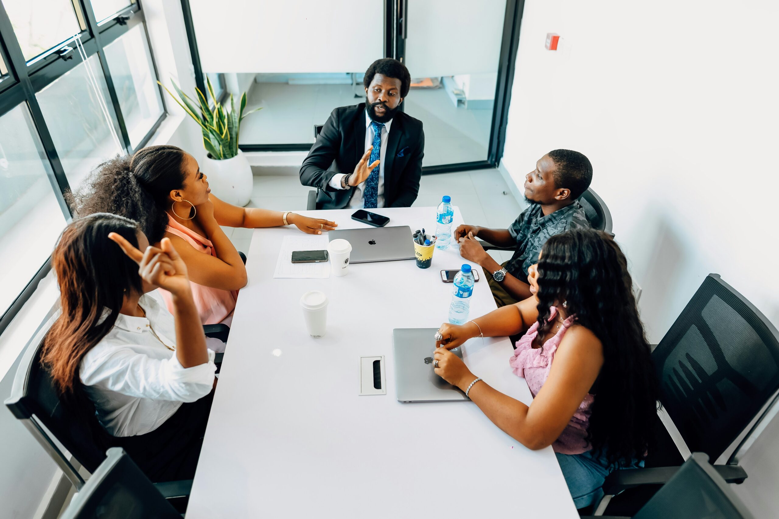 Overhead view of a business meeting in a modern conference room with floor-to-ceiling windows. Four Black professionals are seated around a white table - a man in a dark suit and blue tie gesturing while speaking, and three women in business casual attire listening attentively. The table has laptops, water bottles, smartphones, and other meeting materials scattered on it. The room features contemporary office furniture and a potted plant near the windows.