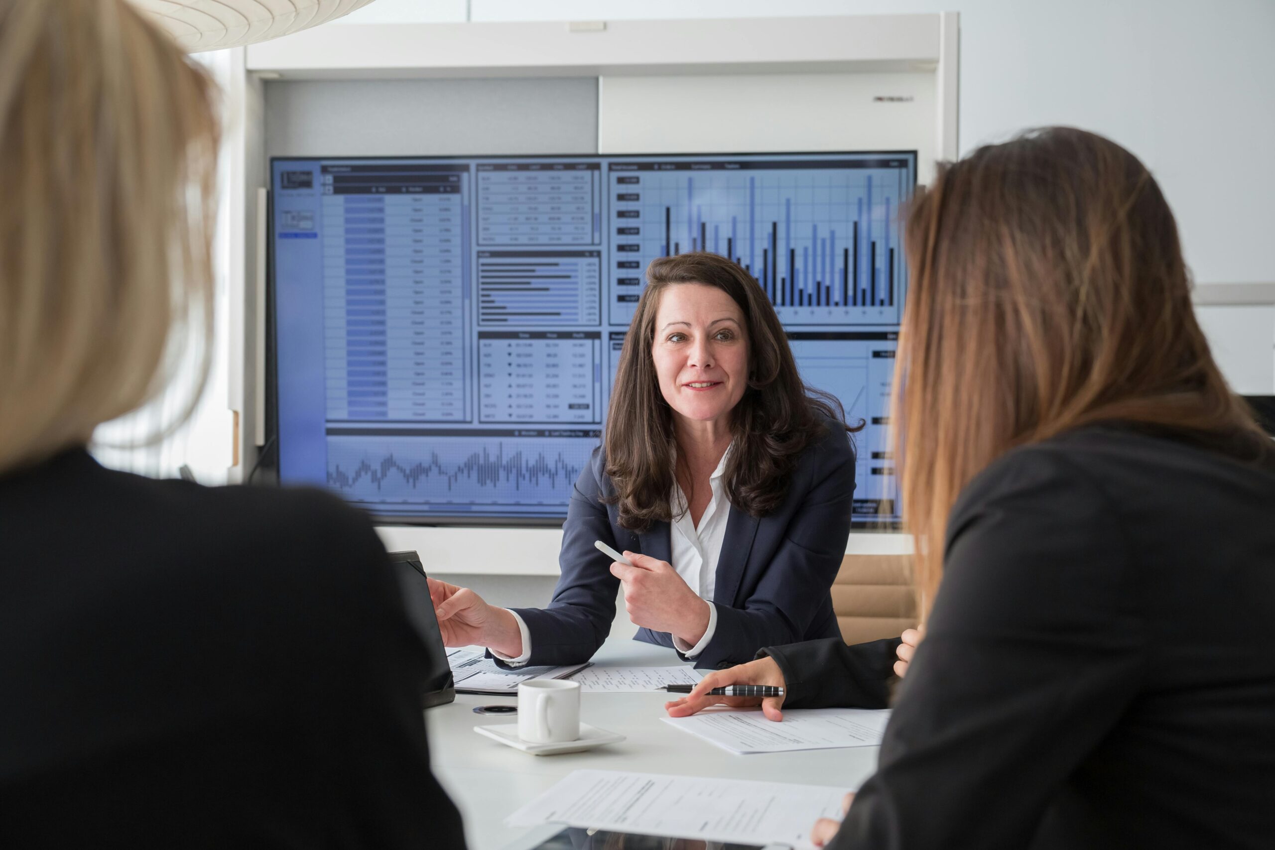 Female executive in business suit presenting data analytics and charts to colleagues in a modern conference room, with multiple dashboard screens displaying financial metrics and graphs in the background