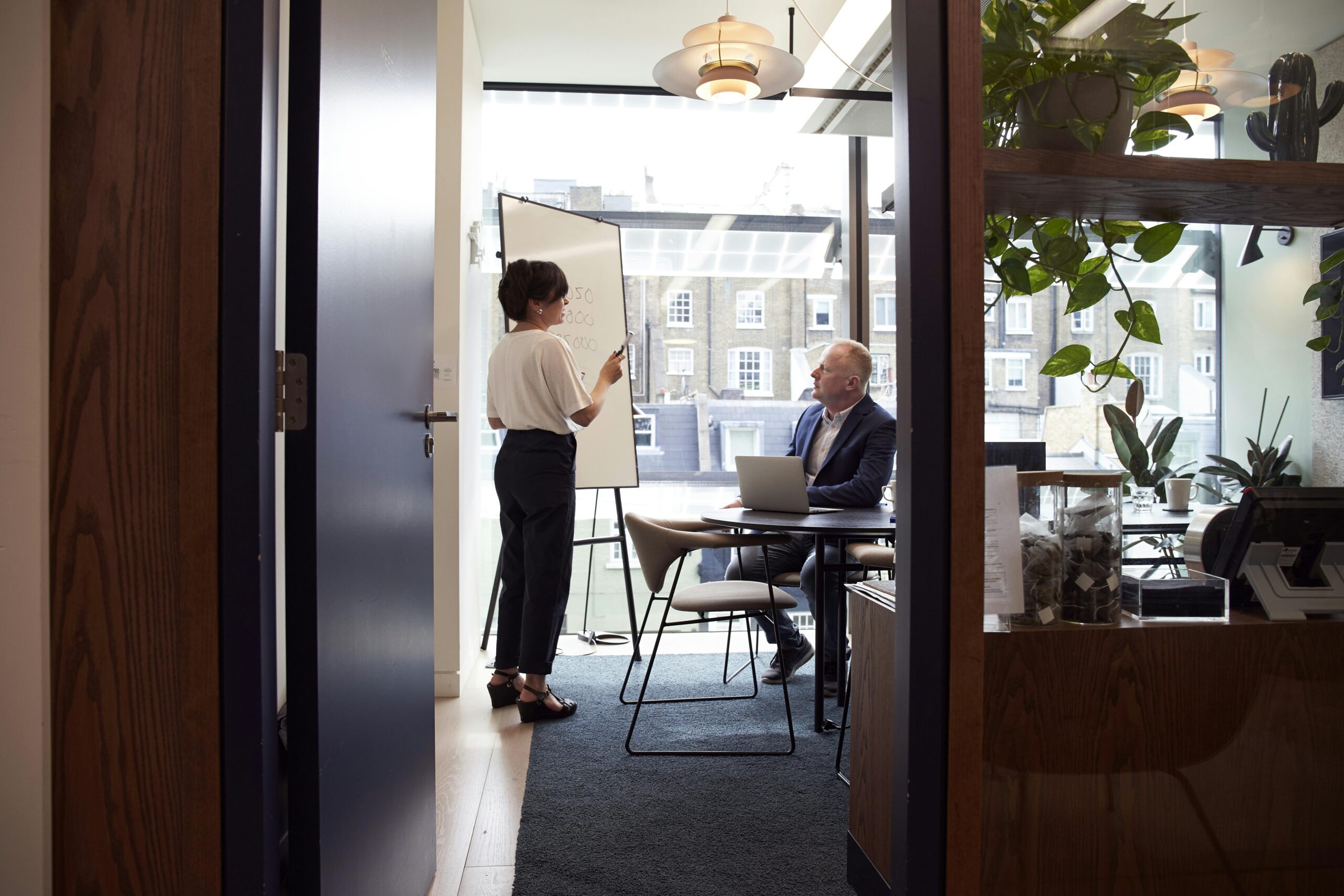 Business professional presenting strategy on whiteboard to executive in modern glass-walled office conference room with city view and contemporary furnishings
